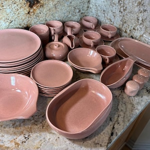 May include: A large collection of dusty rose ceramic dinnerware including stacked dinner plates, small saucers, cups, a lidded sugar bowl, a creamer, oval serving platters, deep bowls, and two small shakers, all featuring a smooth, matte finish.