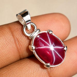 Peut inclure: Un pendentif en argent avec une pierre précieuse rubis étoilé rouge foncé de forme ovale. La pierre en cabochon présente un effet d'étoile à six branches distinct sur sa surface. La pierre est fixée dans une monture en argent à quatre griffes.