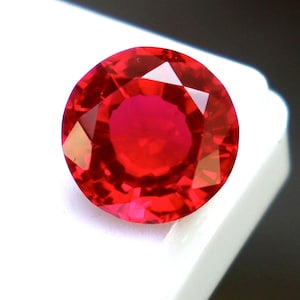 Puede incluir: Una piedra preciosa redonda y facetada de un color rojo intenso y vibrante. La piedra presenta un corte brillante con múltiples facetas triangulares y en forma de cometa que reflejan la luz, creando un aspecto claro y brillante.