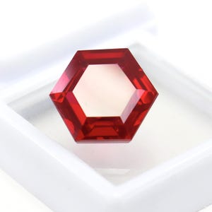 May include: A vibrant red gemstone cut into a precise hexagonal shape with multiple facets that reflect light. The gemstone is displayed inside a small, square white plastic storage box with a clear lid, highlighting its deep crimson colour.