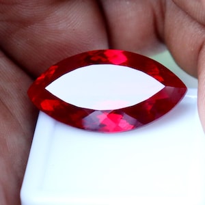 May include: A marquise-cut gemstone with a vibrant, deep red color and a faceted surface that reflects light. The stone is positioned on a small, white, rectangular display base, showcasing its elongated, pointed shape and clear, transparent quality.