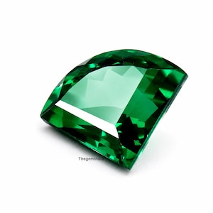 May include: A faceted, fan-shaped gemstone in a vibrant, translucent emerald green color. The stone features precise geometric cuts that create multiple reflective surfaces and sharp edges. The text ThegemIndia is printed in small black letters.