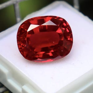 May include: A vibrant, oval-cut red gemstone with a faceted surface that reflects light. The stone displays a deep, saturated crimson hue and is positioned on a white display base, showcasing its clarity and polished, symmetrical edges.