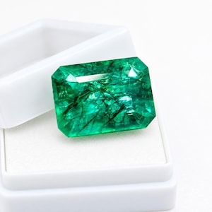 Könnte beinhalten: Ein rechteckiger Smaragd-Edelstein mit leuchtend grüner Farbe und sichtbaren inneren Einschlüssen, der einen facettierten Schliff aufweist, der das Licht reflektiert. Der Edelstein liegt in einer weißen, offenen Schmuckschatulle mit weißem Innenfutter.