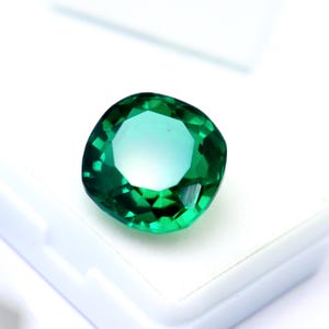 May include: A cushion-cut gemstone with a vibrant, translucent emerald green color. The stone features a faceted surface that reflects light, creating a brilliant appearance with multiple geometric patterns visible across its polished top surface.