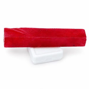 Puede incluir: Una barra de piedra preciosa rectangular de color rojo vibrante con una superficie texturizada y cristalina. La piedra roja alargada está colocada horizontalmente, descansando sobre una pequeña caja de presentación cuadrada de plástico blanco.
