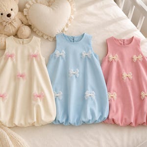 Puede incluir: Tres vestidos sin mangas con corte de globo en color crema, azul claro y rosa. Cada vestido presenta un cuello redondo, una cremallera en la espalda y cinco pequeños lazos decorativos fijados en la tela frontal en un patrón disperso.