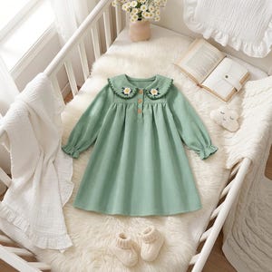 May include: A long-sleeved sage green dress featuring a rounded collar with embroidered white daisy motifs, a button-down placket with three small brown buttons, and ruffled sleeve cuffs. The fabric has a fine vertical ribbed texture.