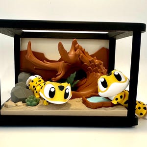 May include: A black-framed display case containing two yellow and black spotted gecko figurines. The scene features a brown sculpted rock formation, grey stones, a small green succulent, and a brown water dish on a tan sandy base.