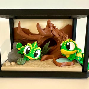 May include: A diorama featuring two green and yellow chameleons inside a black frame. One chameleon sits near a grey rock, while the other is by a small brown water dish. A brown hollow log structure sits in the background on a sandy base.