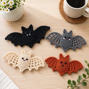 Pode incluir: Quatro ornamentos de morcego em crochê nas cores preto, cinza escuro, creme e laranja queimado. Cada morcego apresenta um padrão de asas detalhado, olhos de contas pretas e expressões faciais bordadas. O morcego preto tem presas brancas.