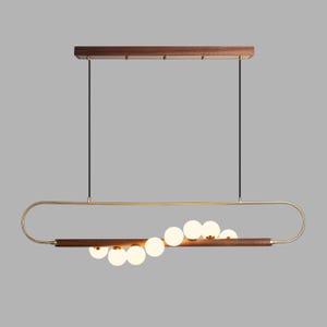 May include: A linear pendant light fixture featuring a horizontal dark wood beam suspended by two thin black cables from a rectangular wooden canopy. A gold-toned metal frame surrounds the beam, which holds eight spherical white glass globes.