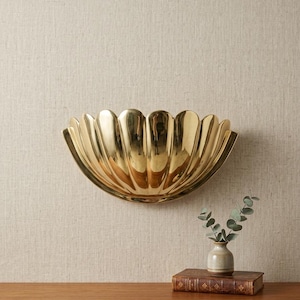 May include: A polished gold-toned metal wall sconce shaped like a scalloped shell. The fixture features a semi-circular design with vertical fluted ridges that create a reflective surface, mounted horizontally to provide an upward light projection.