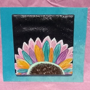 以下が含まれることがあります： 黒い背景にピンク、黄色、紫、ティール色の花びらを持つ様式化されたヒマワリが描かれた正方形のキャンバス画。花の中心部は茶色の質感があり、ティール色のカード台紙に取り付けられています。