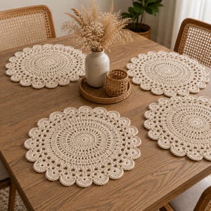 May include: Four round, cream-colored crocheted placemats with a scalloped edge and intricate circular patterns. The placemats are arranged on a wooden table, each featuring a detailed, textured stitch design that radiates from the center outward.