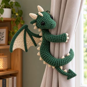 May include: A dark green and cream-colored crochet dragon curtain tieback. The dragon features a textured yarn body, large black eyes, a pair of wings, and a long tail that wraps around a light gray curtain to hold it in place securely.