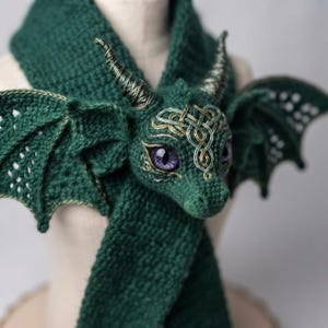 May include: A dark green crochet scarf featuring a three-dimensional dragon head with purple eyes, two horns, and two outstretched wings. The dragon head is decorated with intricate gold-colored embroidery patterns on the forehead and around the eyes.