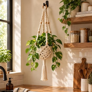 May include: A cream-colored macrame plant hanger featuring a central floral knot pattern, wooden beads, and a long bottom tassel. The hanger holds a green pothos plant in a white pot, suspended from a black wall hook in a kitchen setting.