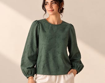 May include: A dark olive green long-sleeve blouse featuring a round neckline, gathered shoulders, and voluminous sleeves with fitted cuffs. The fabric has a subtle, raised, textured pattern throughout the entire garment.