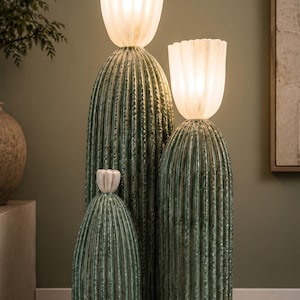 May include: A floor lamp featuring three green, ribbed, cactus-shaped ceramic bases of varying heights. The two tallest bases are topped with illuminated, white, fluted, flower-shaped glass shades, while the shortest base has a small white cap.
