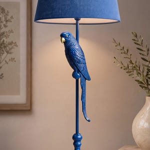 May include: A table lamp featuring a deep blue fabric shade and a matching blue base. The central stem is adorned with a sculpted blue parrot figure perched on a small sphere. The parrot has a gold-colored beak and detailed feather textures.