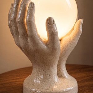 May include: A ceramic table lamp featuring two sculpted hands cupped together to hold a glowing, spherical white glass orb. The base and hands have a speckled, light beige finish with a smooth, glossy texture, creating a warm, diffused light.