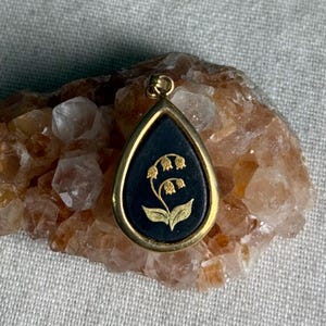 May include: A teardrop-shaped pendant featuring a black inlay with a gold-colored lily of the valley flower design. The pendant is encased in a gold-toned metal frame and includes a small circular bail at the top for attachment to a chain.