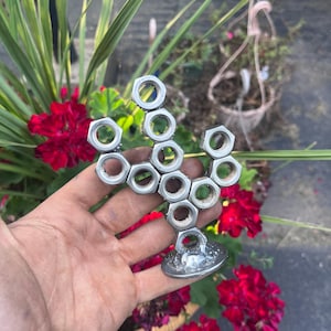 May include: A small sculpture shaped like a cactus, constructed from multiple silver-colored hexagonal metal nuts welded together. The structure features a central vertical column with two branching arms, all mounted on a circular metal base.