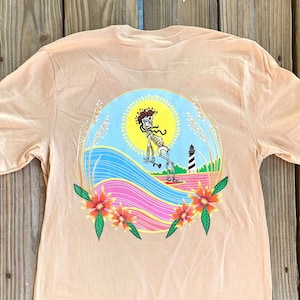 May include: A tan t-shirt with a colorful graphic of a skeleton surfing with a lighthouse in the background. The graphic is surrounded by flowers and a sun.