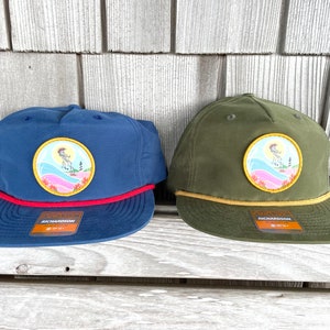 May include: Two baseball caps with a colourful patch featuring a cartoon surfer riding a wave. The cap on the left is navy blue with a red rope trim. The cap on the right is olive green with a yellow rope trim. Both caps have the word "Richardson" on the label.