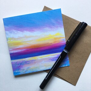 May include: A small square painting depicts a colorful sunset over water, with shades of blue, purple, yellow, and orange. A black Pilot pen and a brown envelope are next to the painting. The painting is on a white surface.