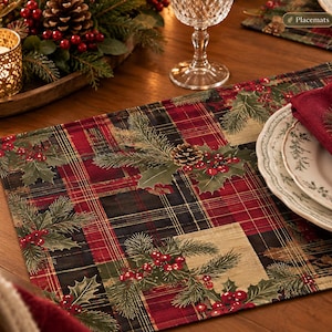 May include: A rectangular fabric placemat featuring a festive pattern of red and black plaid squares interspersed with printed illustrations of green pine branches, brown pinecones, and clusters of bright red holly berries on a dark background.