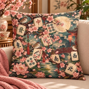 Può includere: Un cuscino quadrato con una stampa raffigurante tessere del mahjong tra fiori di ciliegio rosa, piccoli uccelli, una luna piena, un ponte rosso e una lanterna. Le tessere mostrano vari caratteri cinesi e motivi circolari.