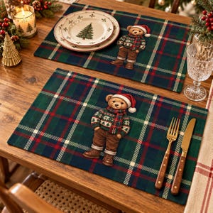 May include: Two rectangular placemats featuring a dark green, navy blue, and red plaid pattern. Each placemat displays a centered illustration of a teddy bear wearing a red Santa hat, a patterned holiday sweater, brown pants, and boots.