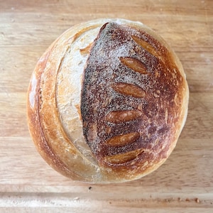 May include: A round loaf of sourdough bread with a dark brown crust and a light brown interior. The bread has a slightly irregular shape and is scored with several lines.