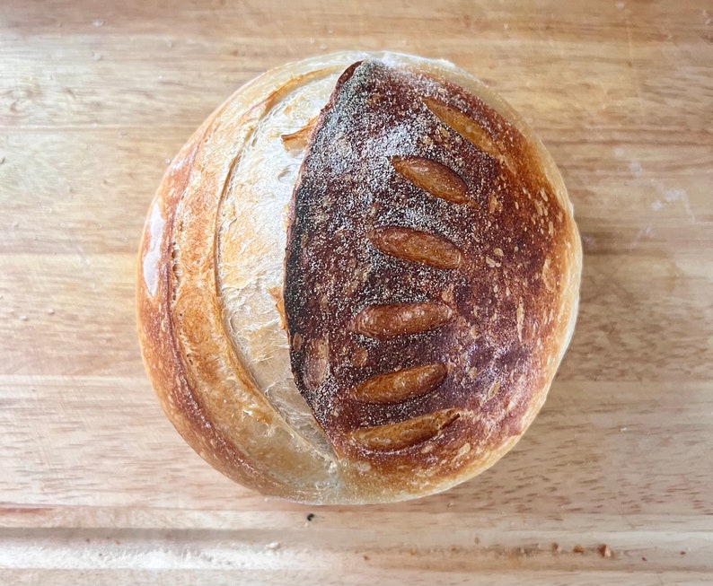 May include: A round loaf of sourdough bread with a dark brown crust and a light brown interior. The bread has a slightly irregular shape and is scored with several lines.