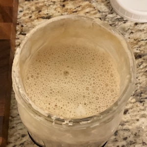 May include: A glass jar filled with a light brown liquid with a foamy texture. The liquid is bubbling and has a light brown color.