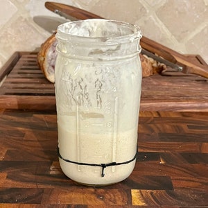 May include: A clear glass jar with a black rubber band around the top. The jar is filled with a light brown liquid and has markings on the side indicating ounces.