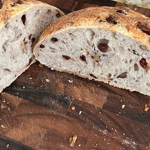 May include: A loaf of sourdough bread sliced in half, revealing a light brown crumb with dried cranberries and nuts. The bread is on a wooden cutting board.