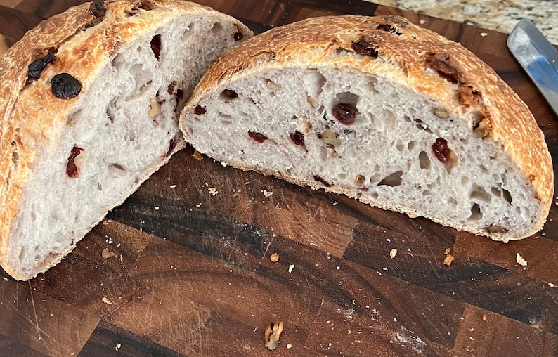 May include: A loaf of sourdough bread sliced in half, revealing a light brown crumb with dried cranberries and nuts. The bread is on a wooden cutting board.