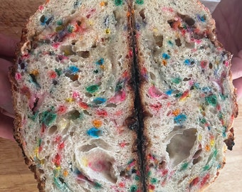 May include: A loaf of sourdough bread sliced in half, revealing a white crumb speckled with colorful sprinkles. The bread has a slightly browned crust.