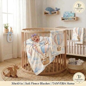 May include: A soft fleece blanket measuring 30x40 inches featuring a light blue and cream pattern of hot air balloons carrying rabbits, clouds, and stars. The blanket is draped over a round wooden crib containing a baby with a purple bow.