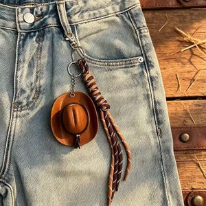 May include: A keychain featuring a miniature brown cowboy hat charm and a braided cord made of dark brown and tan rope with white flecks. The keychain is attached to a silver-toned metal ring and lobster clasp, hanging from a belt loop of blue jeans.