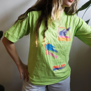 May include: A lime green t-shirt with a colorful graphic design featuring a blue and purple flag with the text "Bugle Boy" and "Sunrise Sun" printed on the front.