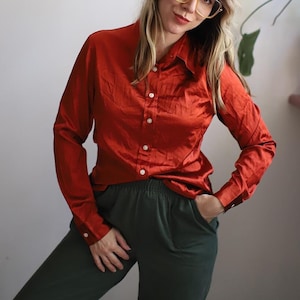 Puede incluir: Una mujer con una camisa roja de satén con botones y pantalones verde oscuro. Lleva gafas y tiene la mano en el bolsillo.