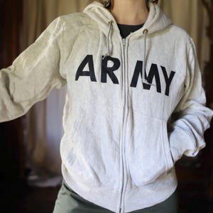 Może przedstawiać: Jasnoszara bluza z kapturem zapinana na zamek z napisem "ARMY" wydrukowanym pogrubionymi czarnymi literami na piersi. Bluza posiada kaptur ze sznurkiem, zamek błyskawiczny na całej długości i dwie przednie kieszenie.