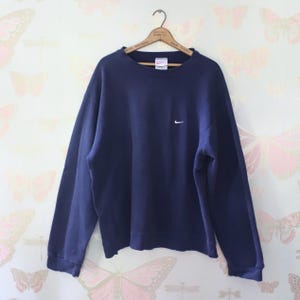 Puede incluir: Una sudadera Nike azul marino con un peque&ntilde;o logotipo blanco de Nike en el pecho. La sudadera de manga larga y cuello redondo est&aacute; colgada de una percha de madera. El fondo tiene un estampado de papel pintado con mariposas.