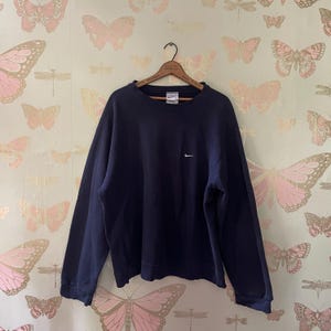 Puede incluir: Una sudadera azul marino de cuello redondo con un peque&ntilde;o logotipo blanco de Nike en el pecho. La sudadera est&aacute; colgada en una percha de madera sobre un fondo de pantalla con mariposas rosas.