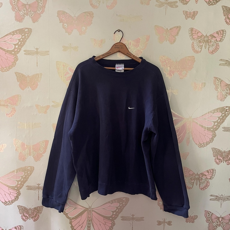 Puede incluir: Una sudadera azul marino de cuello redondo con un peque&ntilde;o logotipo blanco de Nike en el pecho. La sudadera est&aacute; colgada en una percha de madera sobre un fondo de pantalla con mariposas rosas.