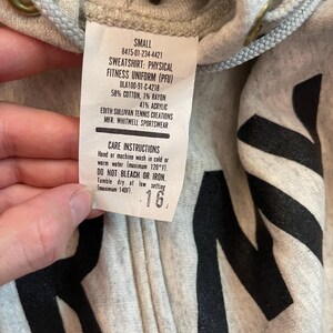 Może przedstawiać: Jasnoszara bluza z kapturem zapinana na zamek z czarnymi literami. Etykieta głosi "SWEATSHIRT: PHYSICAL FITNESS UNIFORM (PFU)" i zawiera instrukcje pielęgnacji. Bluza jest oznaczona jako "SMALL" i ma kaptur ze sznurkiem.
